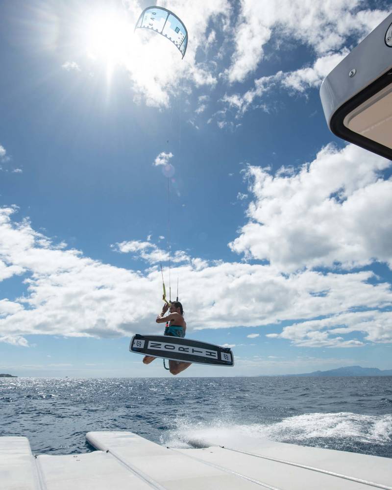A kiteboarder soars through the air above the sparkling blue ocean, propelled by a large kite against a backdrop of clouds and sun.