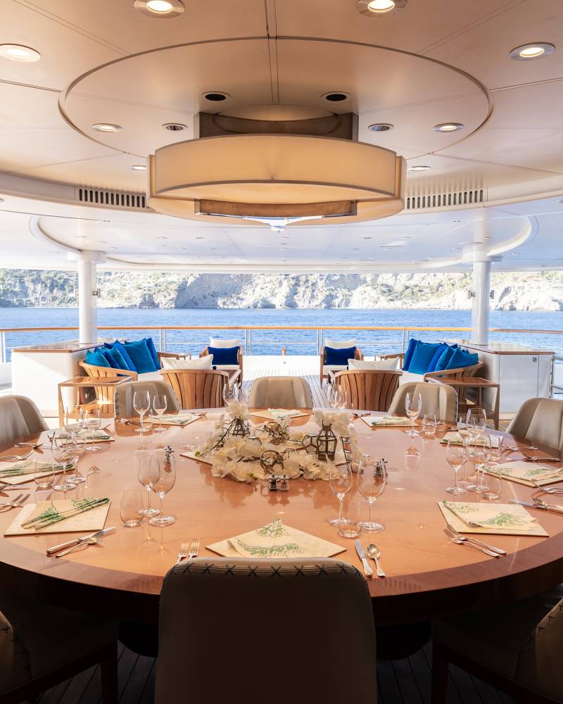 A beautifully set dining table on a yacht's deck offers a stunning view of the Mediterranean Sea and rocky coastline.