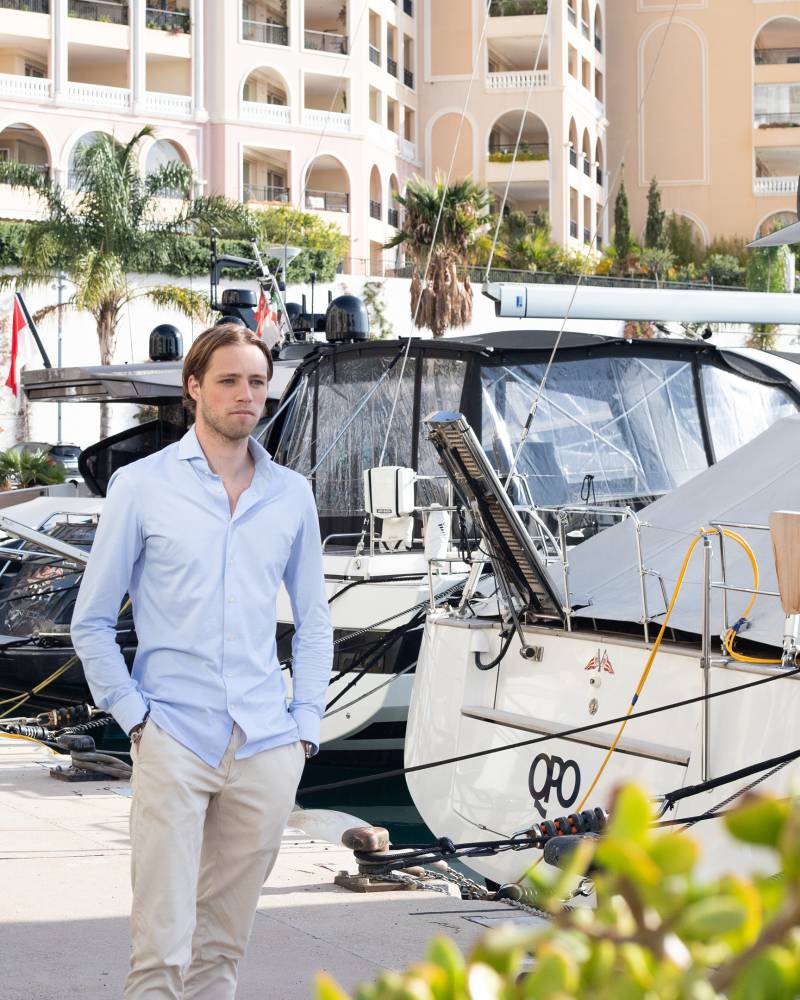 A man in a light blue shirt and beige pants stands on a dock in front of several yachts, with a cactus in the foreground and a modern building in the background.