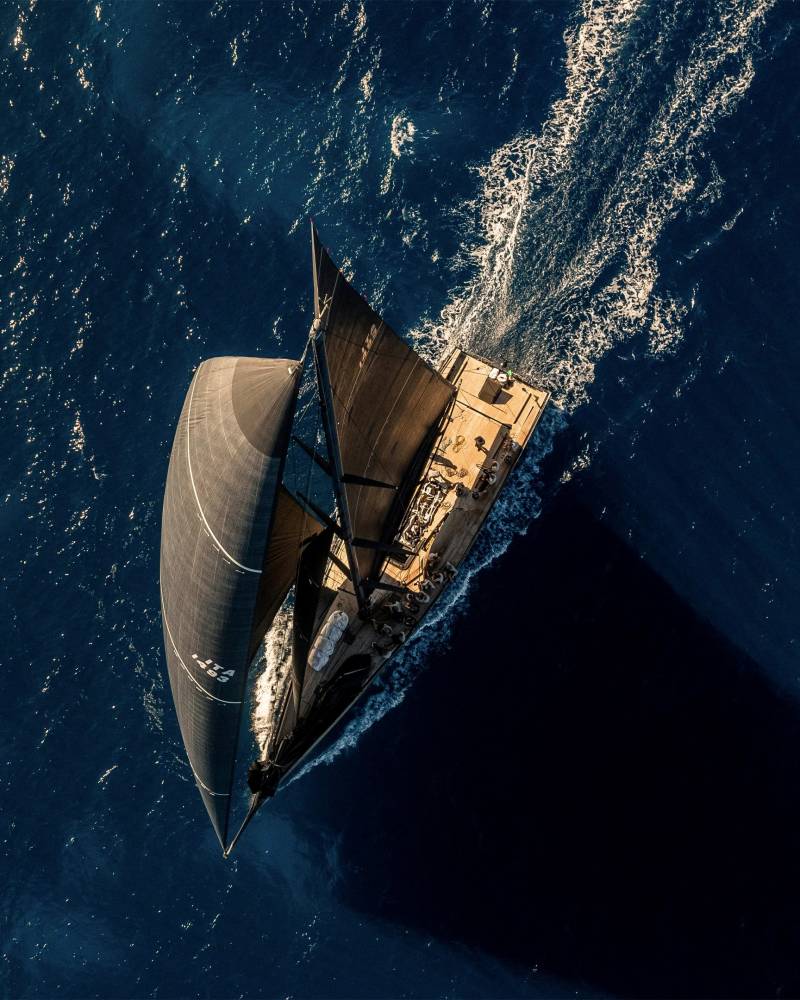 A sleek sailboat cuts through the deep blue ocean, its dark sails billowing in the wind.