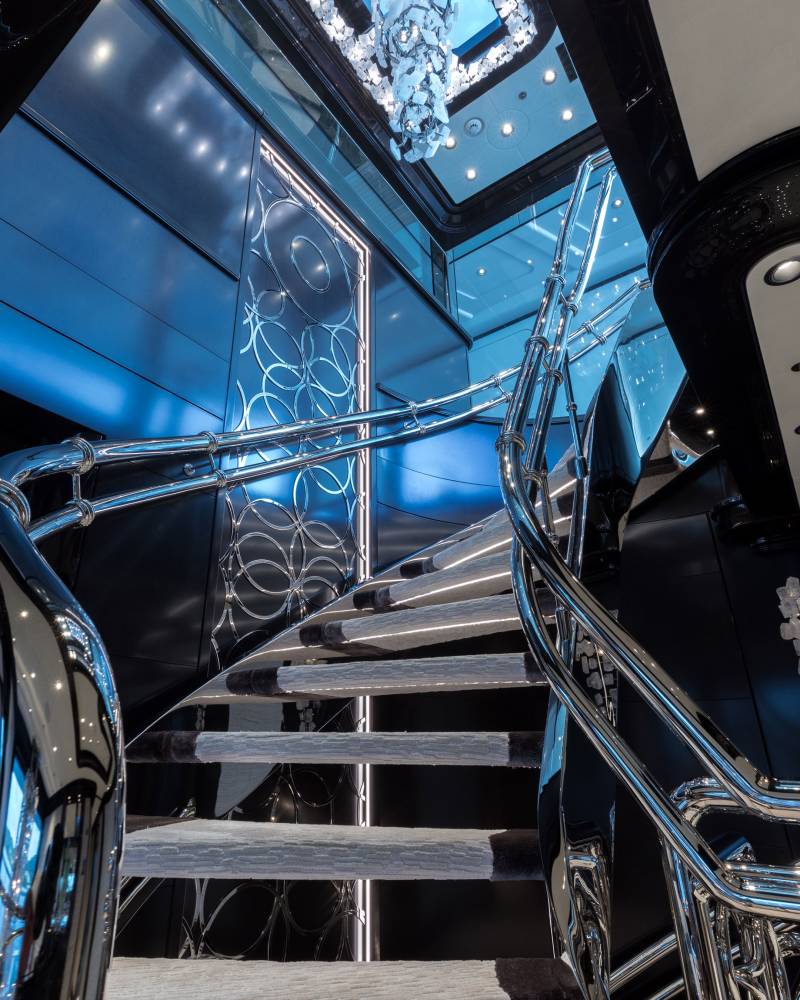 A luxurious spiral staircase with plush carpeting and a dazzling chandelier ascends into a bright, modern space.