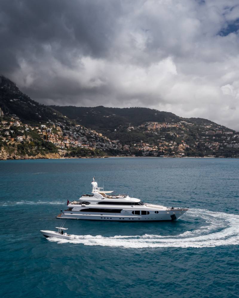 A luxurious white yacht cruises through the turquoise waters, leaving a swirling wake near a densely populated, verdant coastline under dramatic clouds.