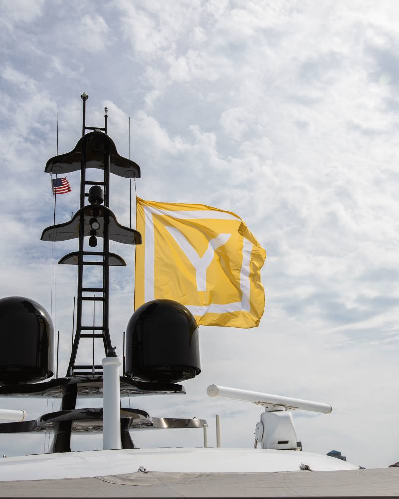 A large yellow flag with a white "Y" billows in the wind above a yacht's radar array against a cloudy sky.