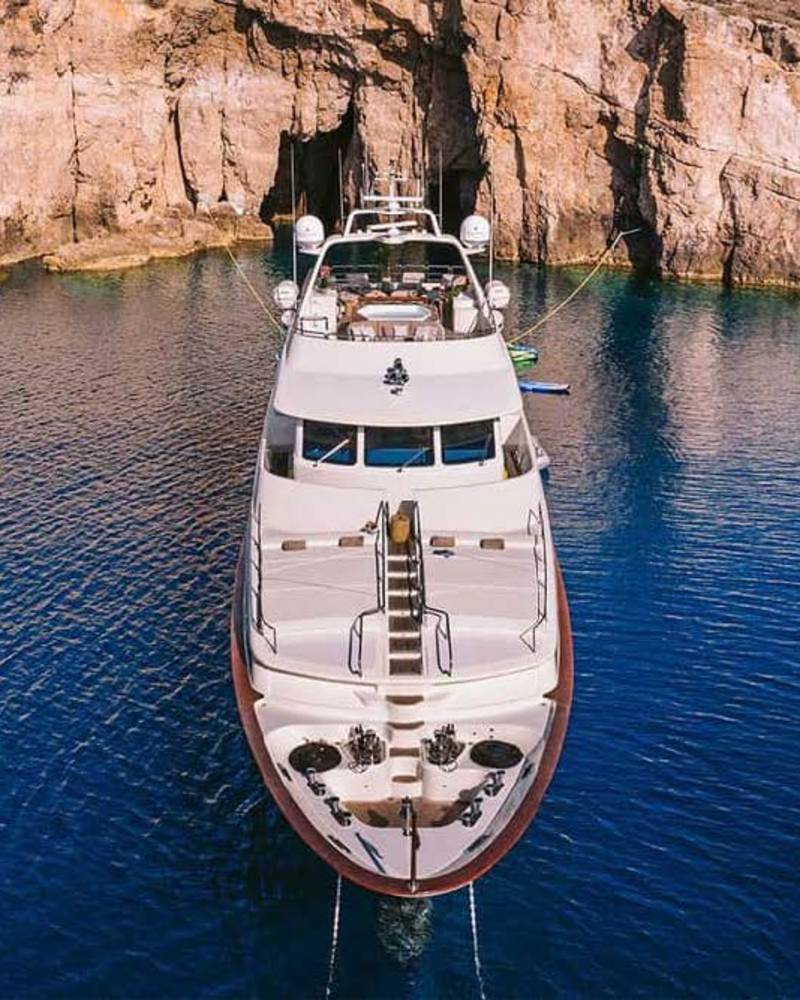 A luxurious white yacht is anchored in the crystal-clear blue waters of a secluded cove framed by rugged cliffs.