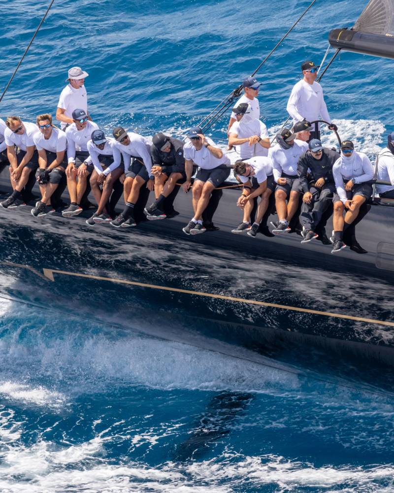 A team of sailors leans over the dark hull of a racing yacht as it cuts through the choppy blue ocean.