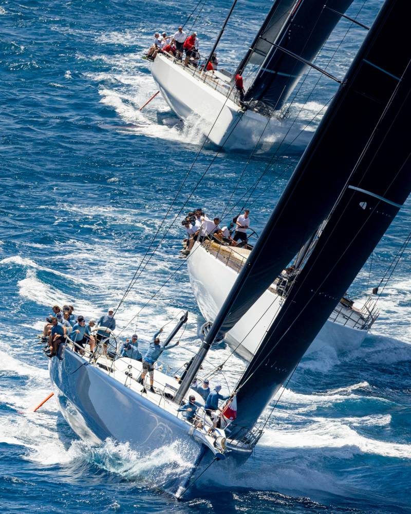 Two sleek sailboats with dark sails race through choppy blue waters under a bright sun.