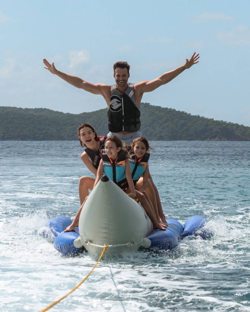 A family enjoys a thrilling ride on a banana boat, pulled across the sparkling blue ocean under a clear sky.
