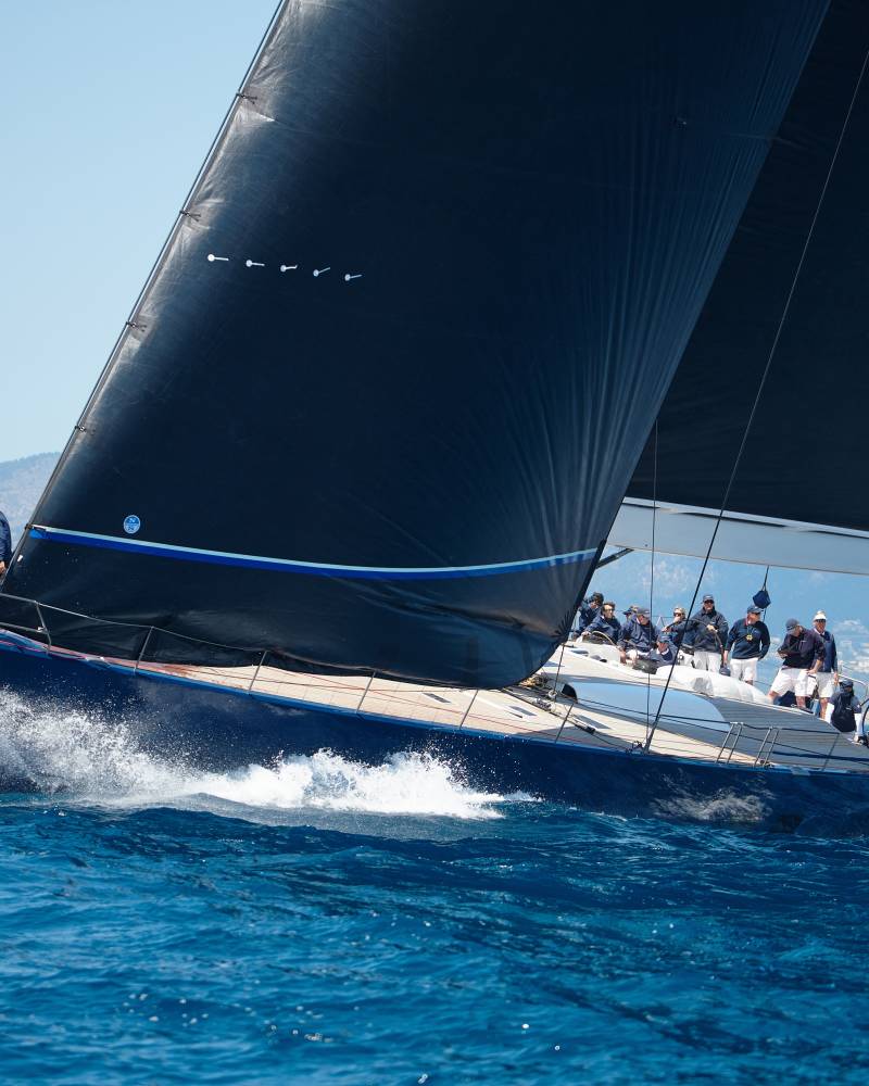 A large, sleek sailing yacht cutting through the deep blue waters of the ocean, with passengers on board enjoying the ride against a backdrop of clear skies and distant mountains.