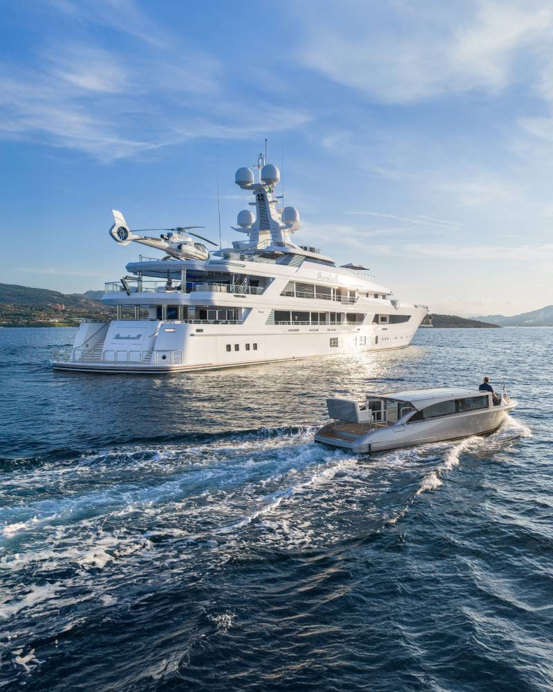 A sleek superyacht with a helicopter on deck cruises through the sparkling blue water, followed by a smaller tender boat.