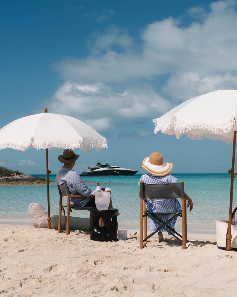 Two people relax under beach umbrellas on a pristine white sand beach, gazing out at a turquoise sea and a distant yacht.