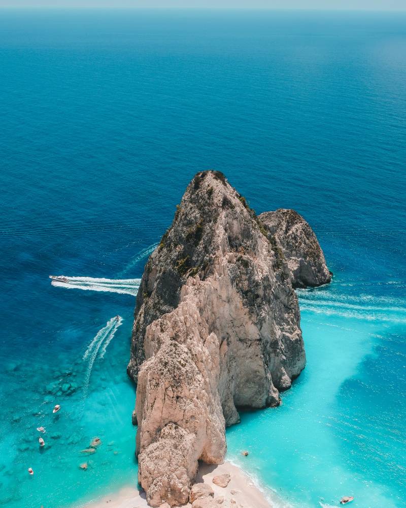 Azure waters surround a dramatic rocky island and a pristine white sand beach dotted with small boats.