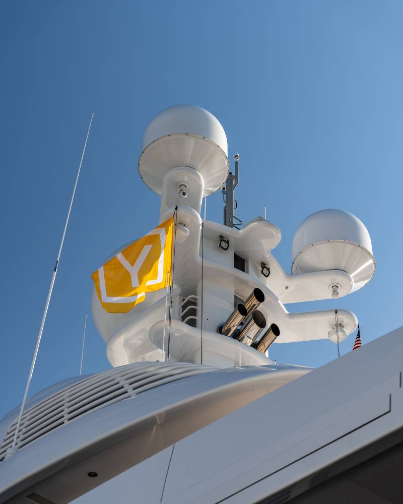 A bright yellow flag with a white "Y" flutters from a luxury yacht's mast against a clear blue sky.