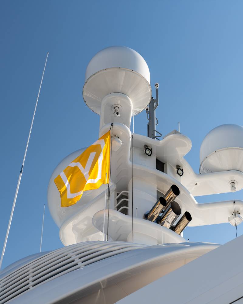 A bright yellow flag with white geometric symbols flutters against a clear blue sky atop a sleek white yacht.