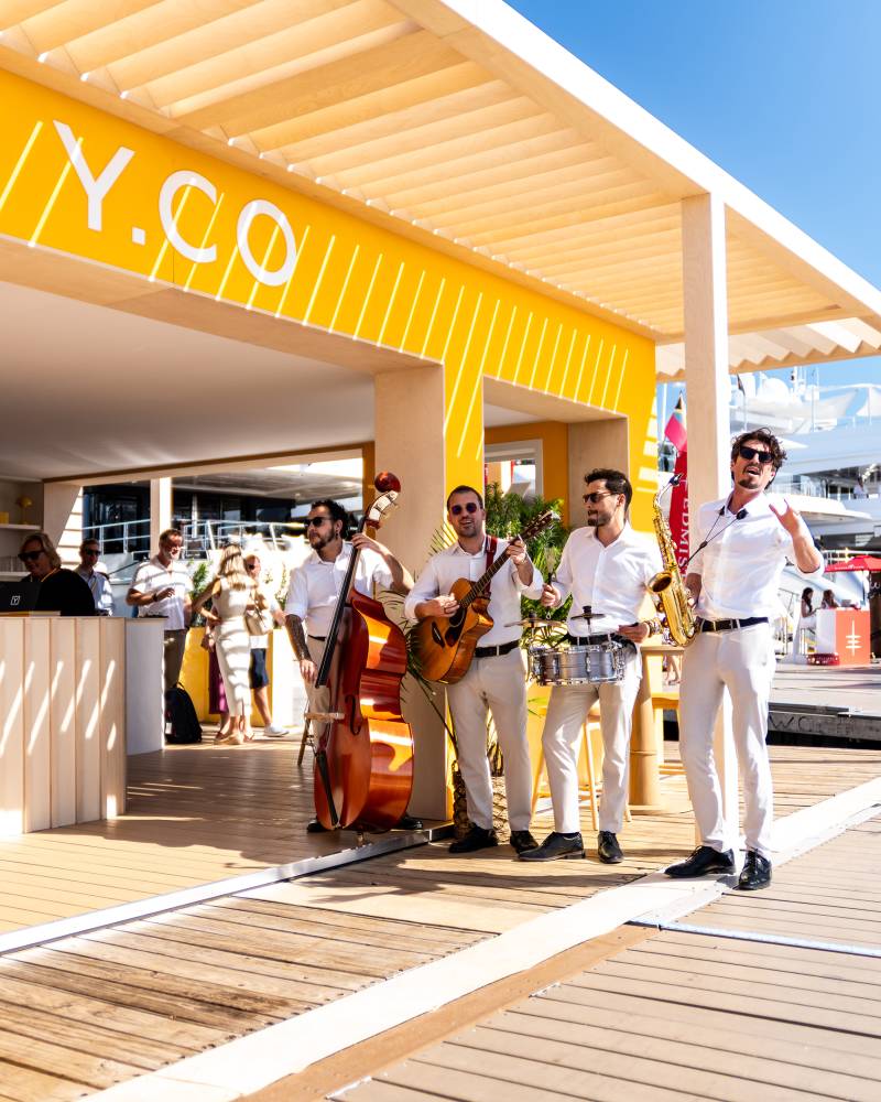 A jazz band plays on a sunny boardwalk in front of a bright yellow building with yachts in the background.