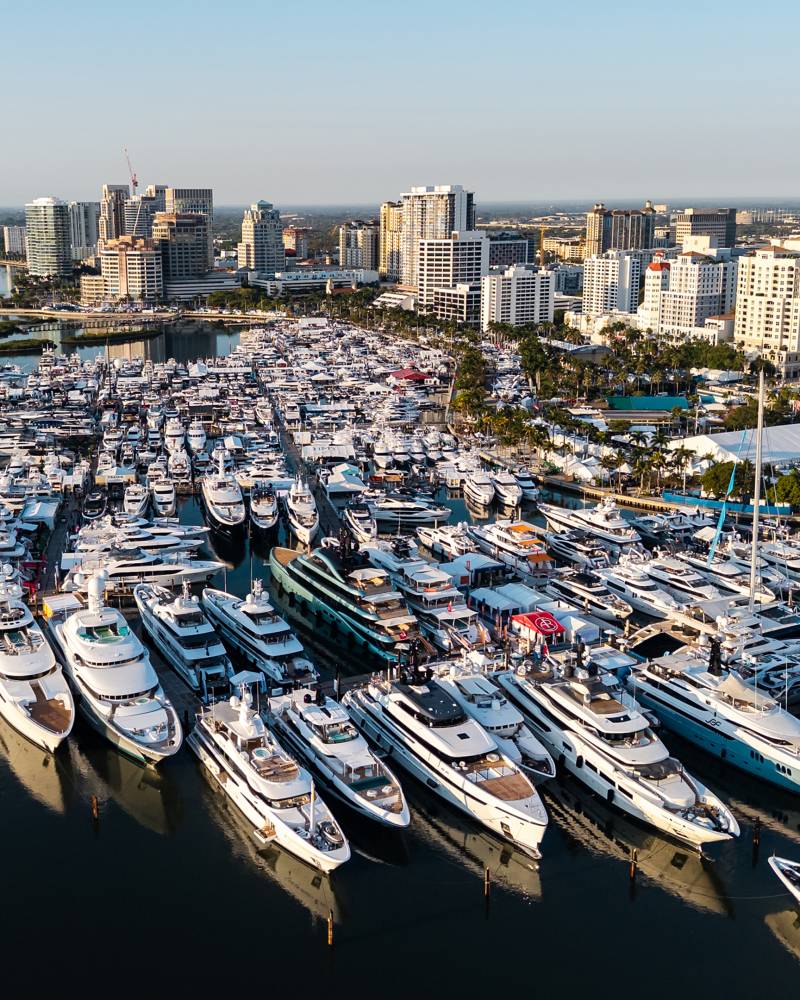 Aerial view of numerous yachts docked at the Palm Beach International Boat Show with a city skyline in the background.