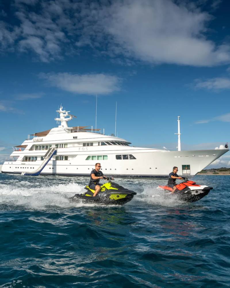 A large, luxurious yacht is anchored in the distance, while two people on jet skis are riding through the waves in the foreground against a backdrop of a bright blue sky with fluffy white clouds.