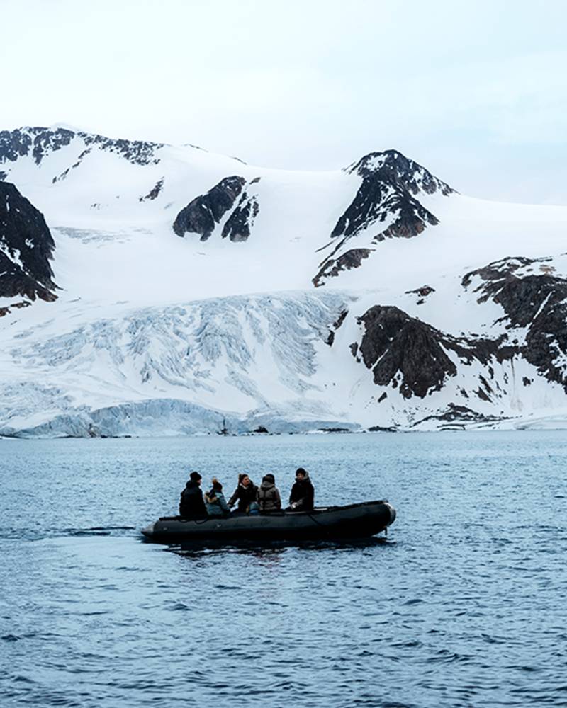 A small boat carrying several people floats on the calm waters of a fjord, surrounded by towering snow-capped mountains in the background.