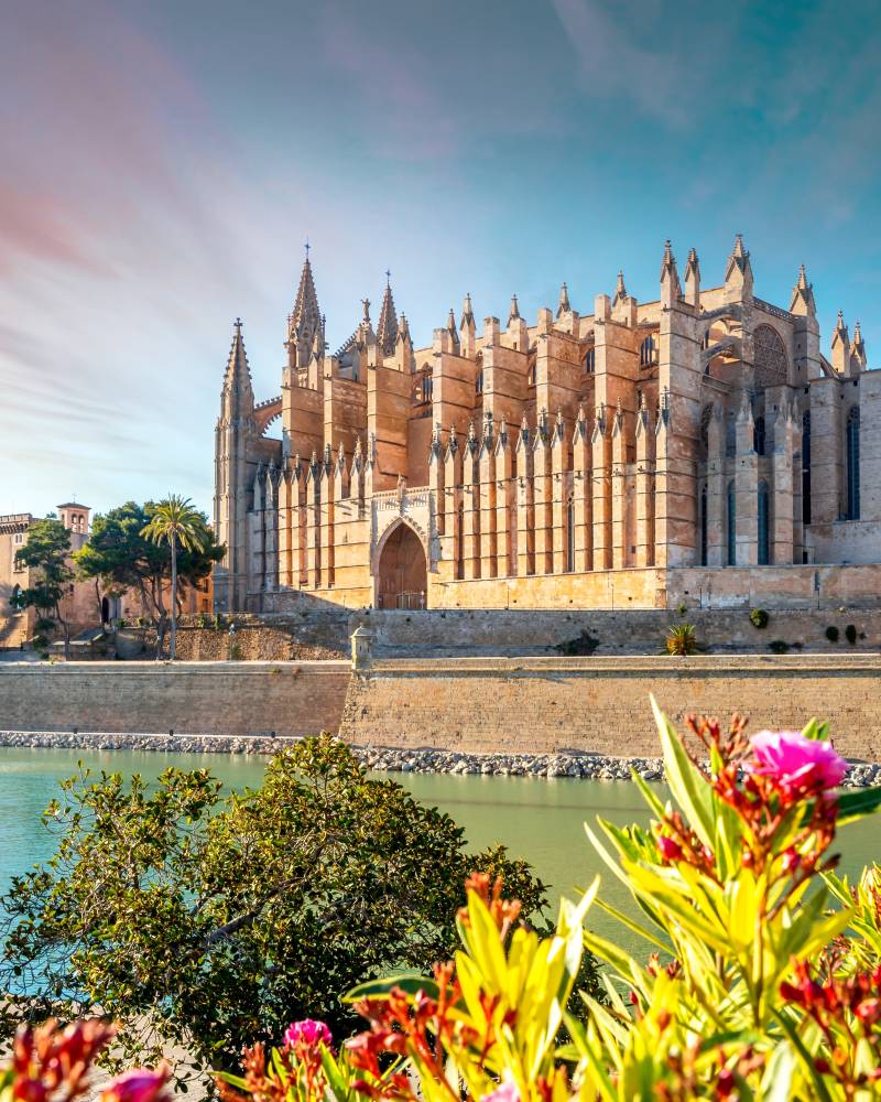A magnificent cathedral with ornate Gothic architecture stands tall against a vibrant blue sky, surrounded by a tranquil body of water and lush, colorful foliage in the foreground.