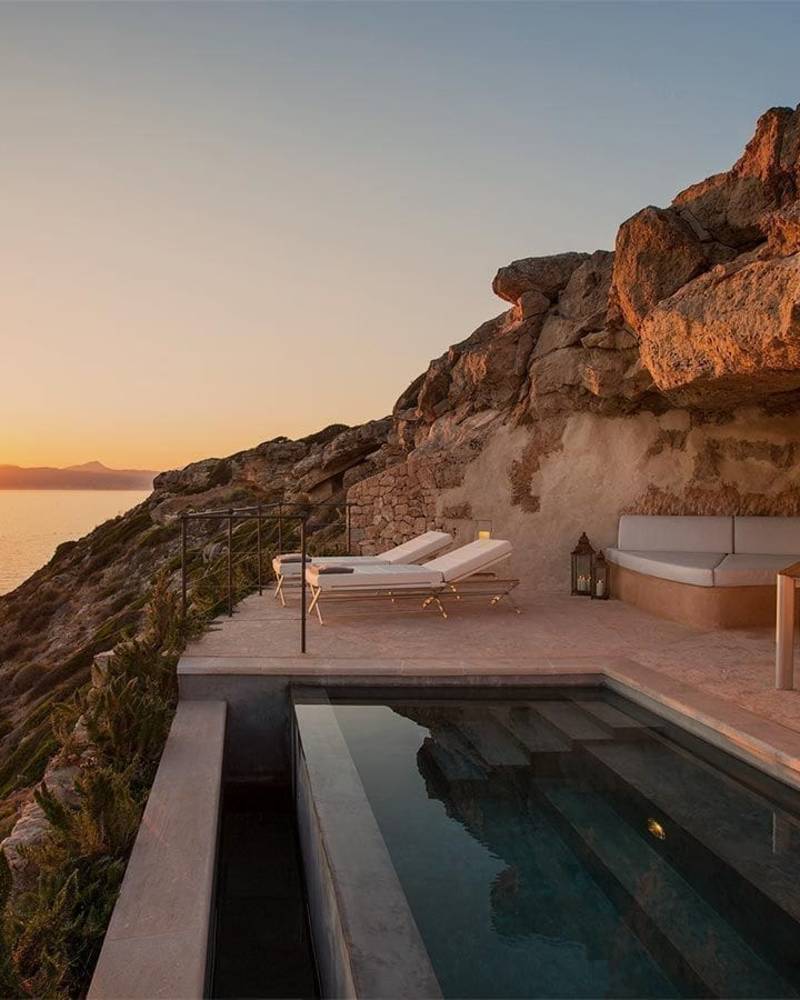A stunning cliffside villa with a private pool overlooking a serene body of water, bathed in the warm glow of a breathtaking sunset.