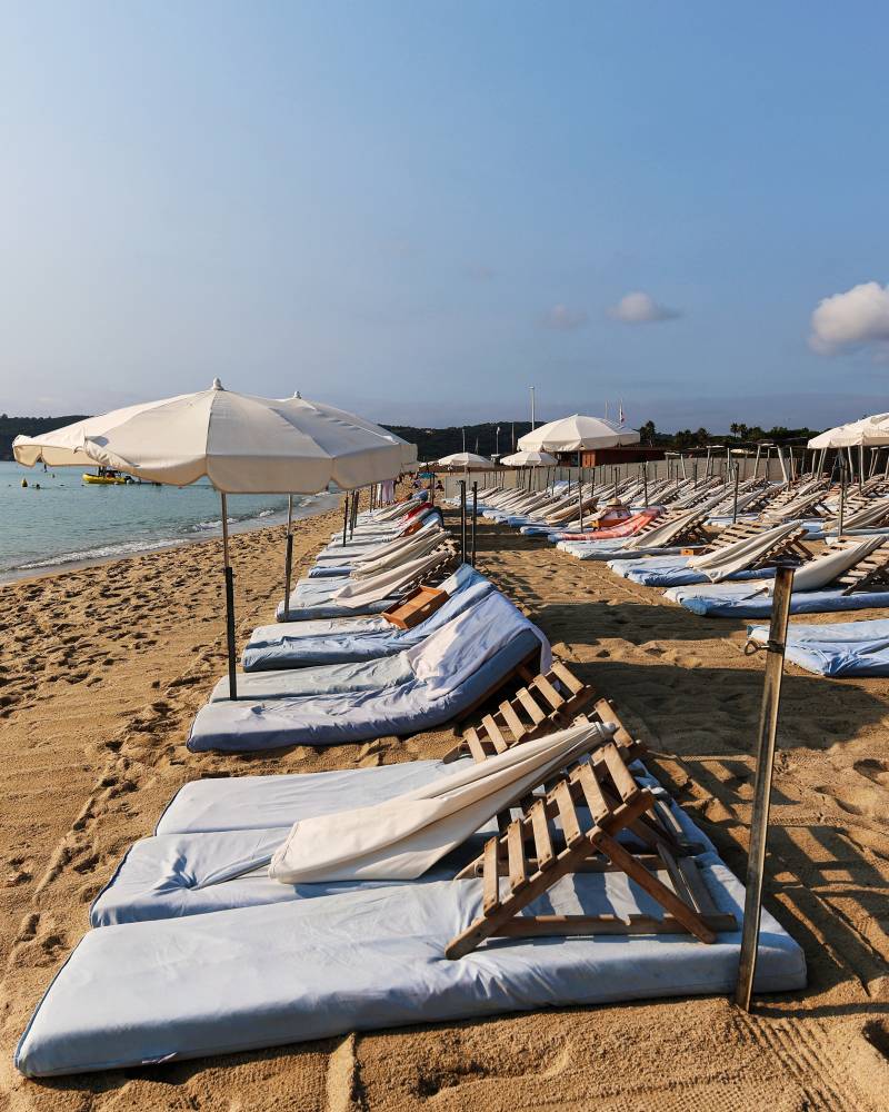A wooden pier extends into the clear turquoise waters of a sunny beach, leading towards a distant, tree-covered coastline.
