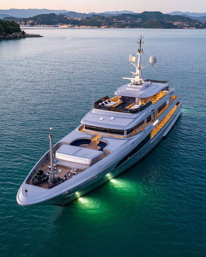 A large, luxurious yacht with a sleek, modern design is seen floating on a calm, blue-green sea, surrounded by a picturesque coastal landscape in the background.