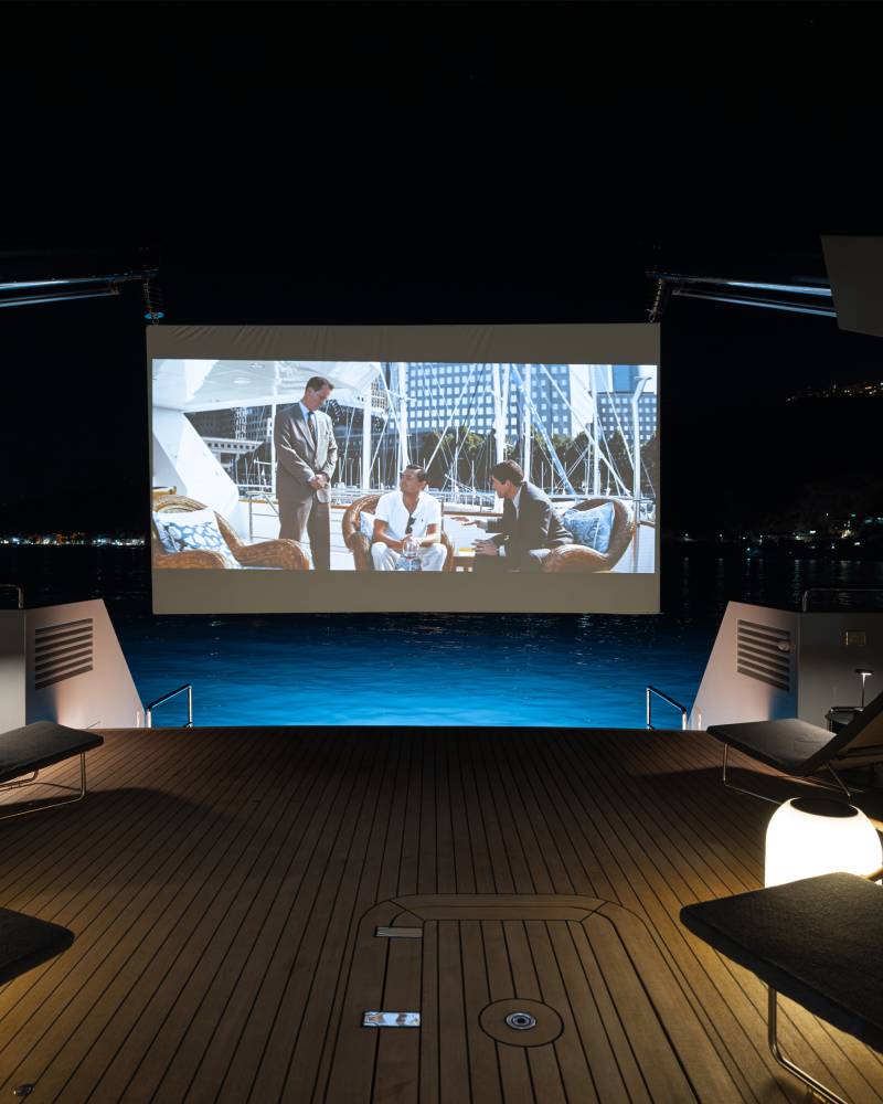 A luxurious outdoor cinema setup with comfortable lounge chairs, a large projection screen, and a scenic backdrop of a city skyline at night.