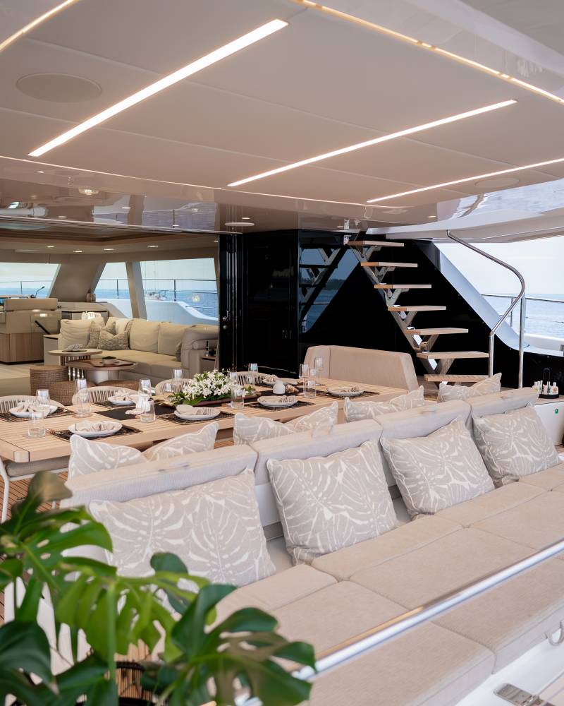 A luxurious outdoor dining area with plush seating and a table set for a meal is visible on a yacht's deck, with the ocean and a distant coastline in the background.