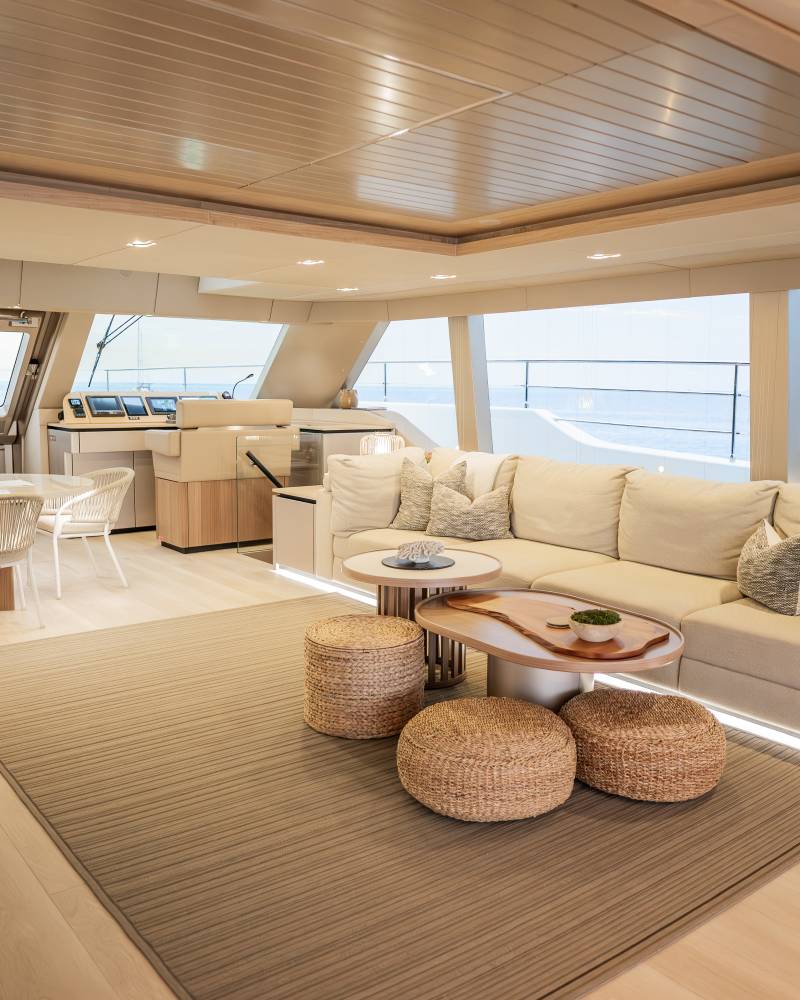 A spacious yacht interior features a large, comfortable sectional sofa and woven ottomans in the foreground, with a dining area and expansive windows overlooking the ocean in the background.