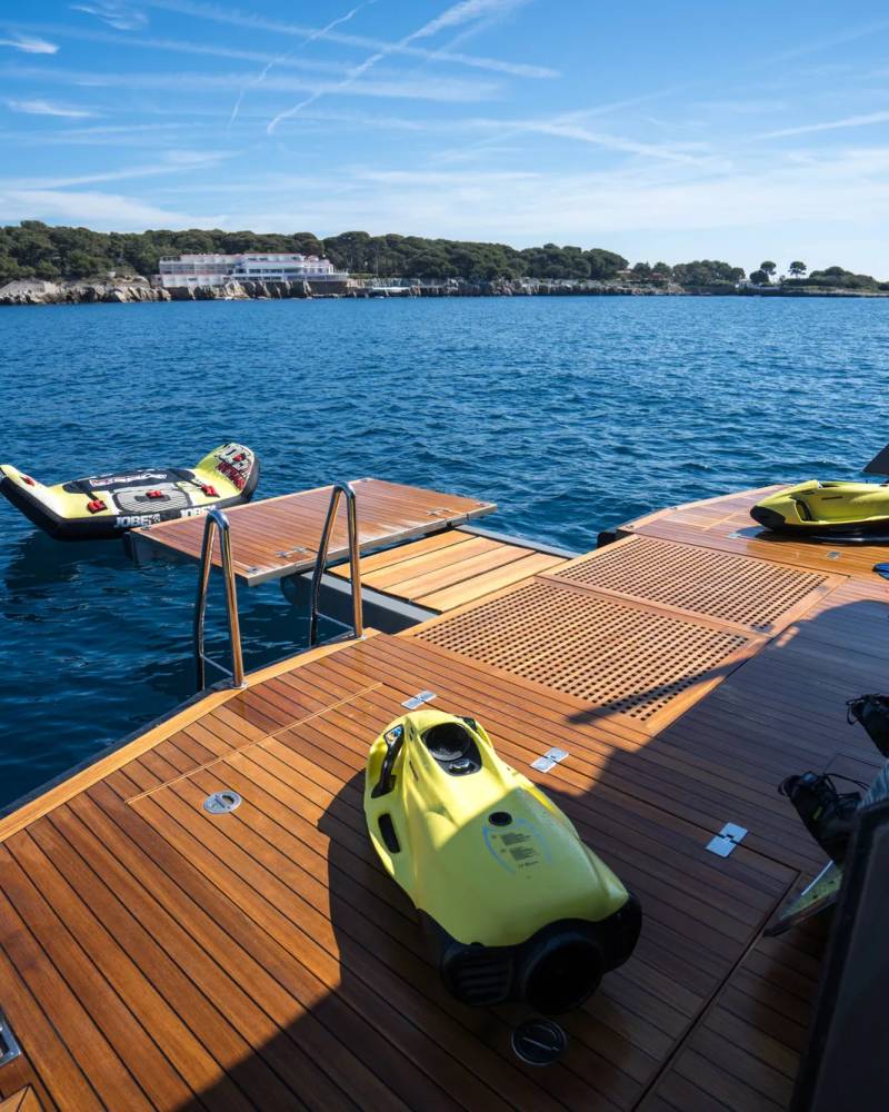 Two yellow underwater scooters rest on the wooden deck of a yacht, with a floating inflatable mat and a ladder extending into the calm blue water, and a tree-lined coast with buildings in the distance.