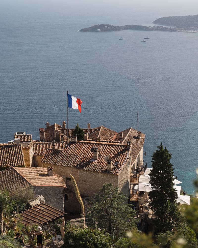 A French flag flies over a village with terracotta roofs in the foreground, overlooking a sparkling blue bay and distant coastline in the background.