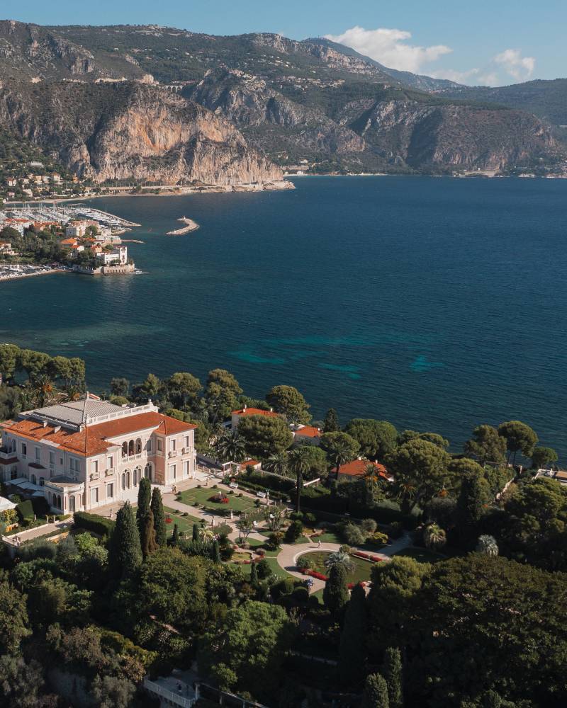 A grand villa with manicured gardens sits nestled amongst lush greenery in the foreground, overlooking a sparkling blue bay dotted with sailboats and a coastal town nestled against dramatic cliffs in the background.