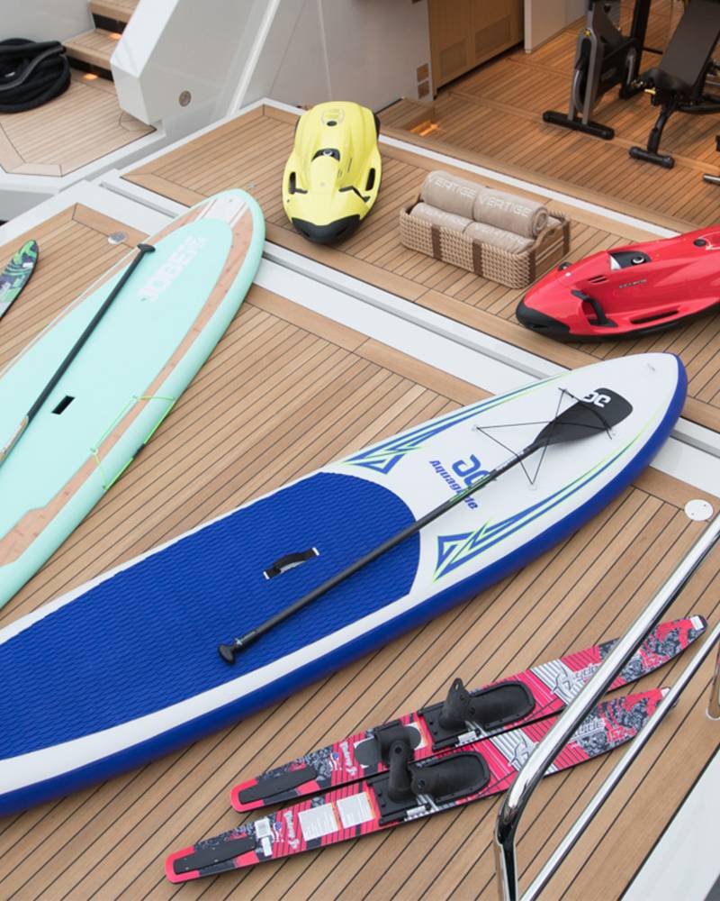 In the foreground, paddleboards and skis rest on a yacht's deck, while the background reveals exercise equipment and watercraft.