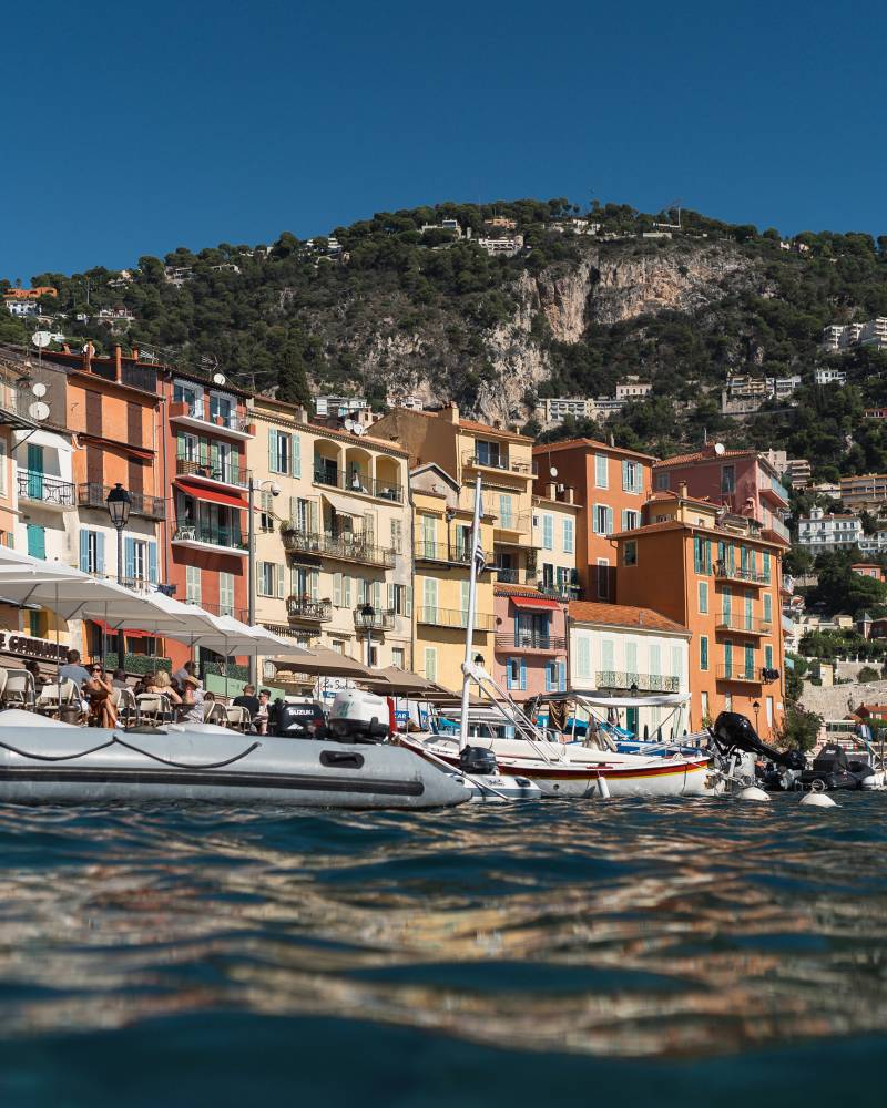 In the foreground, boats bob on the water, while the background features a colorful row of buildings nestled against a tree-covered hillside under a clear blue sky.