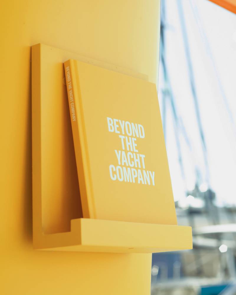 A yellow book titled "BEYOND THE YACHT COMPANY" rests on a shelf against a bright yellow wall, with a blurred background of sailboats.