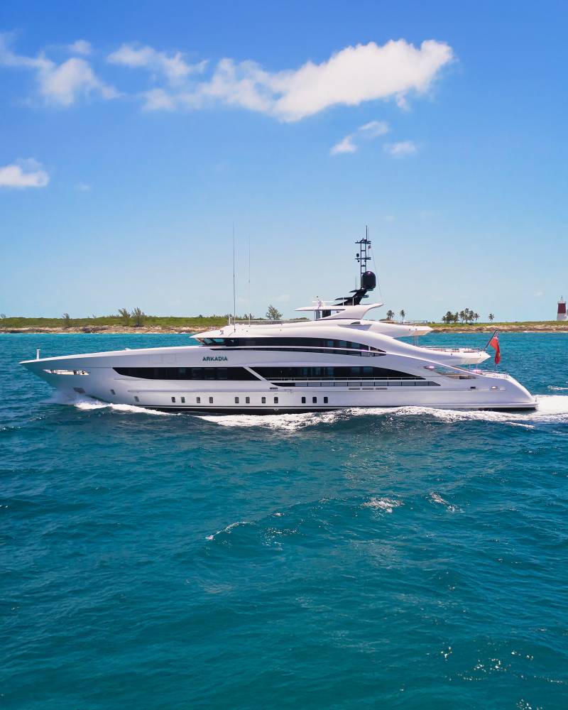 A sleek white yacht cruises through turquoise waters with a sandy coastline and blue sky in the background.