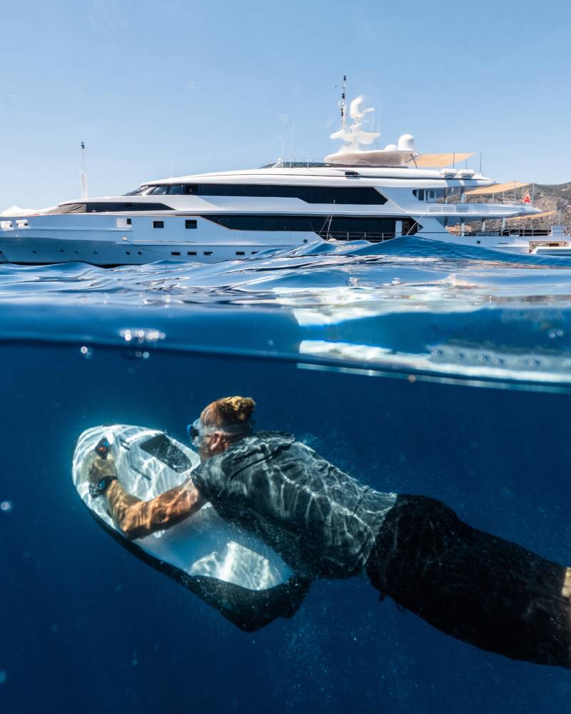 A person glides underwater with a sea scooter in the foreground, while a large white yacht is anchored in the background near a sun-drenched coastline.