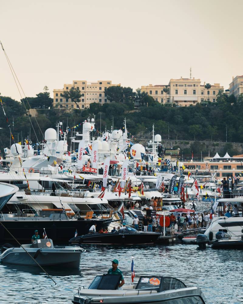 Several small boats are in the foreground on the water, with a large fleet of yachts and buildings on a hill in the background.