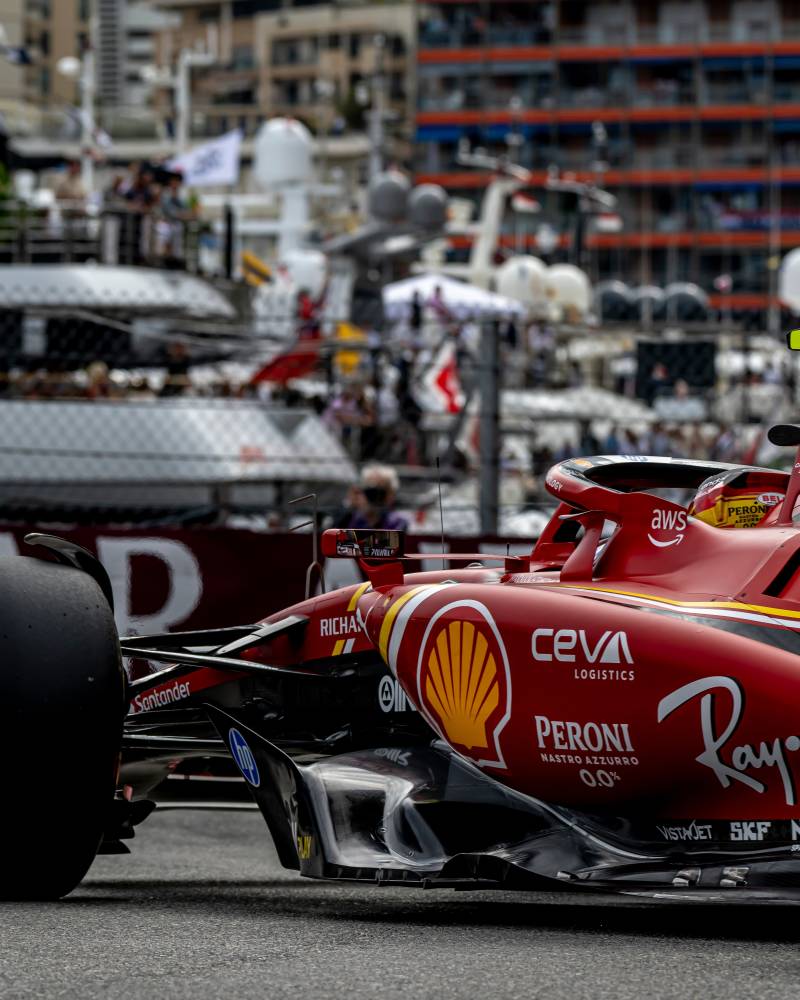 A red Formula 1 race car speeds along a track with grandstands and buildings in the background.