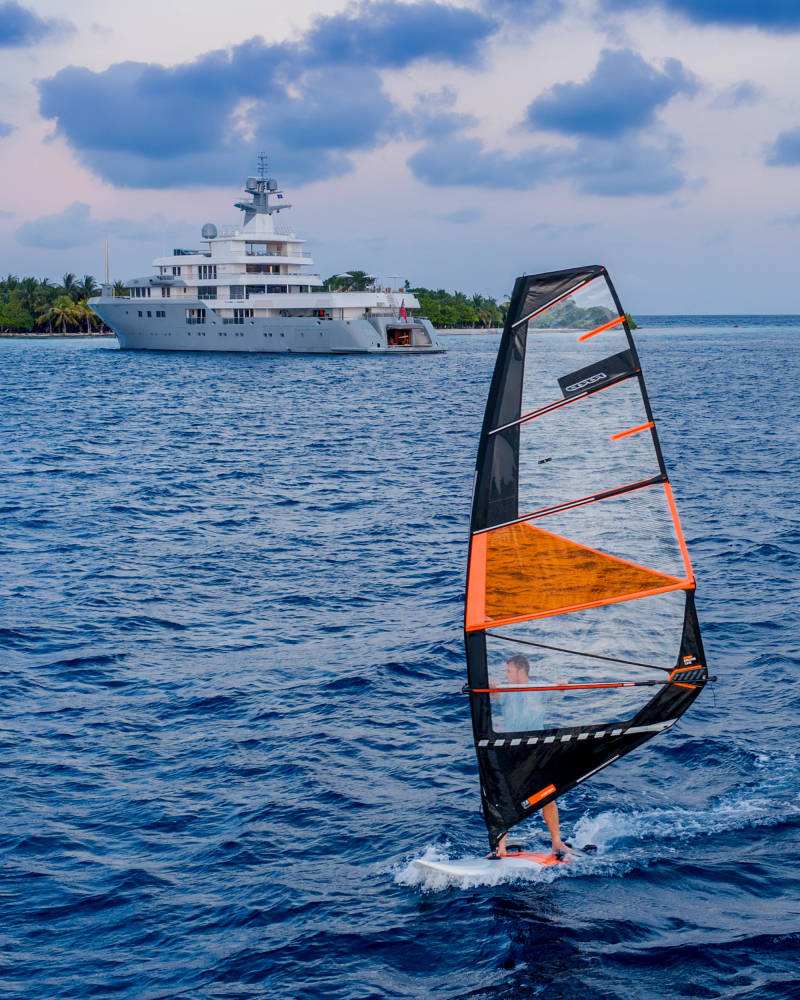 A person windsurfing in the foreground sails past a large yacht anchored near a tropical island in the background.