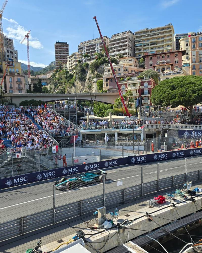 A race car speeds down a track in front of a grandstand filled with spectators, with a backdrop of densely packed buildings and cranes on a sunny day.