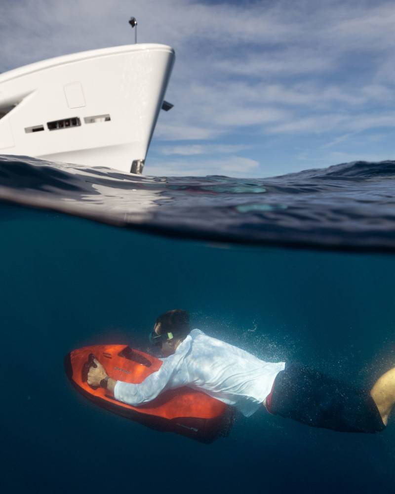 A man in a suit swims underwater on a red scooter, with a large white yacht sailing above him in the blue ocean.