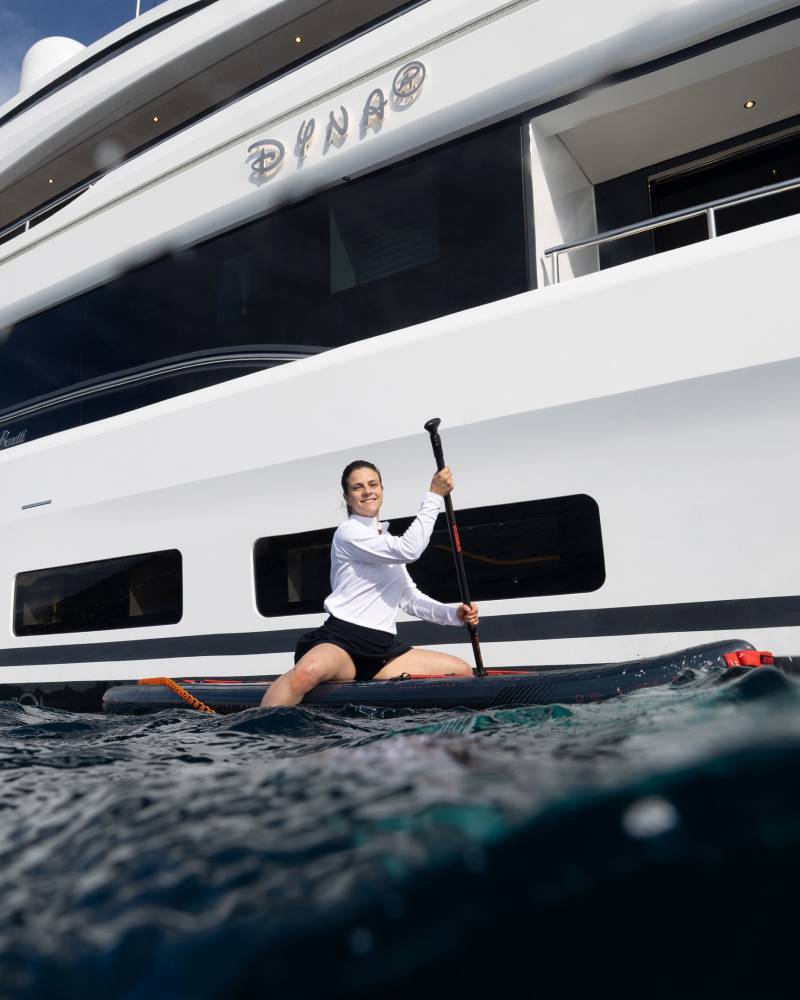 A woman paddles a stand-up paddleboard in the foreground, with a large white yacht named "Dyna" in the background.