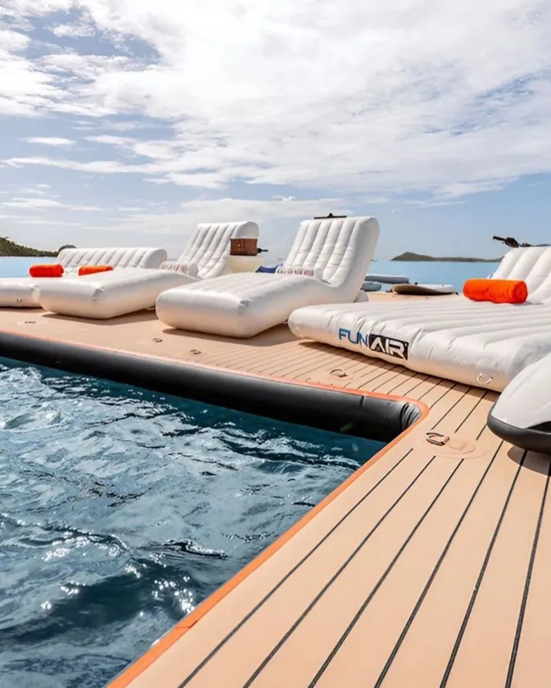 Inflatable loungers and a jet ski rest on a floating dock in the foreground, with a calm blue ocean and lush green islands in the background.