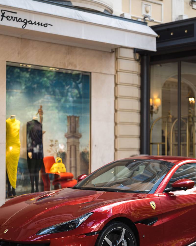 A red Ferrari is parked in front of a Salvatore Ferragamo store with doormen standing by the entrance.