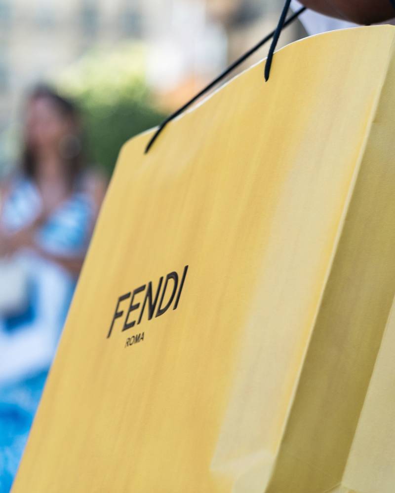 A close-up of a Fendi shopping bag is held by a person in the foreground, with two blurred figures in the background.