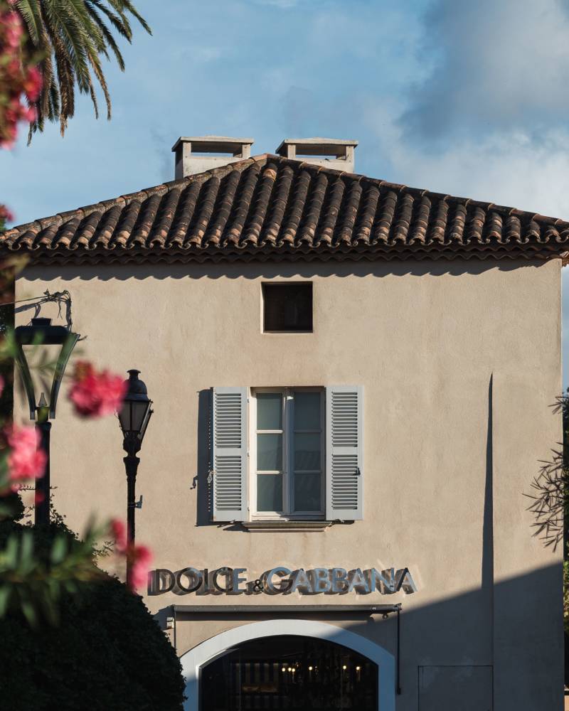 Pink flowers frame a Dolce & Gabbana store with a tiled roof and a single window, set against a bright sky with scattered clouds.