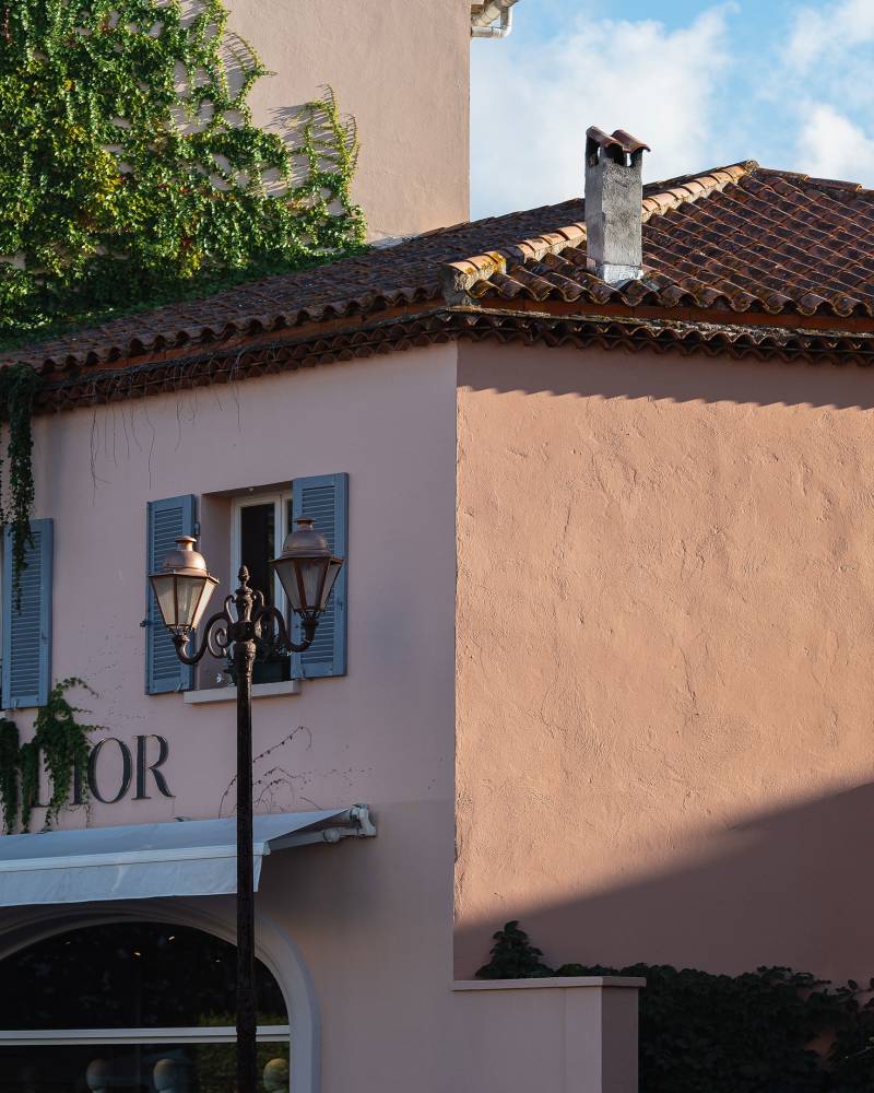 A pink building with blue shutters and a Dior sign in the foreground is set against a background of green foliage and a tiled roof under a cloudy sky.
