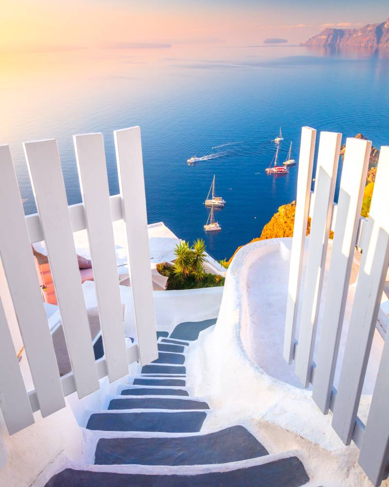A white picket fence and stone steps lead down to a vibrant blue sea dotted with sailboats, with a hazy, sunlit coastline in the distance.