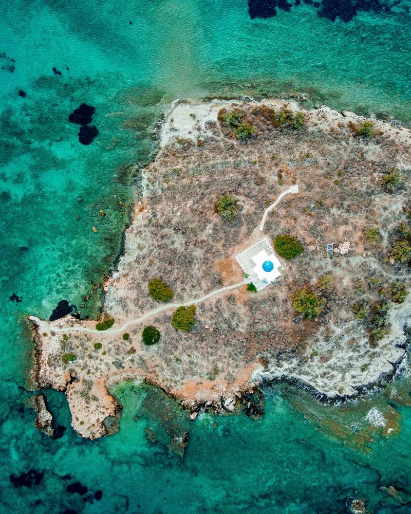 A small, arid island with a white-domed building sits in the vibrant turquoise waters of the ocean.