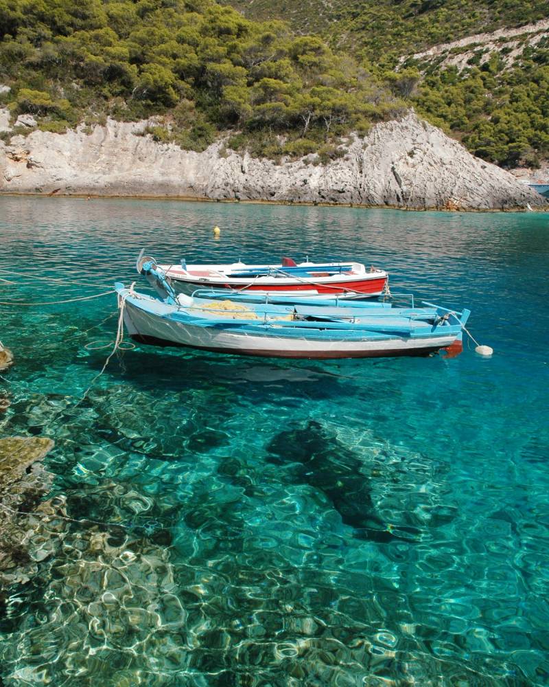 Two small boats float on crystal-clear turquoise water in the foreground, with a rocky, tree-covered hillside rising in the background.